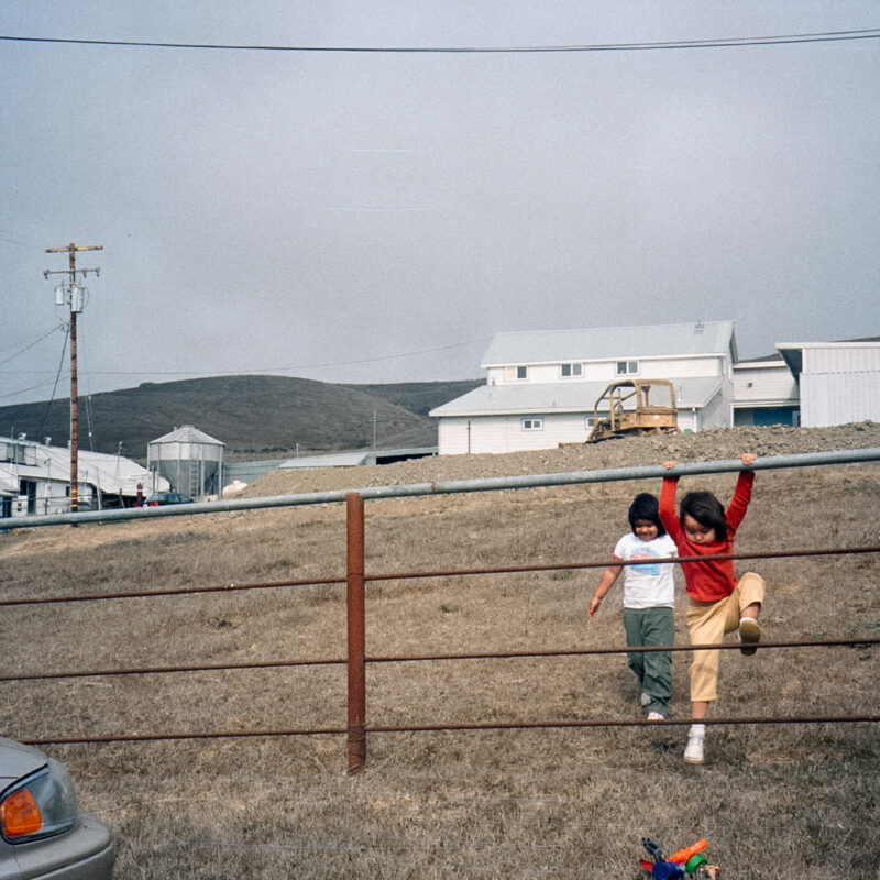 Two children playing in a fence in a field with white barn-style structures in the background.